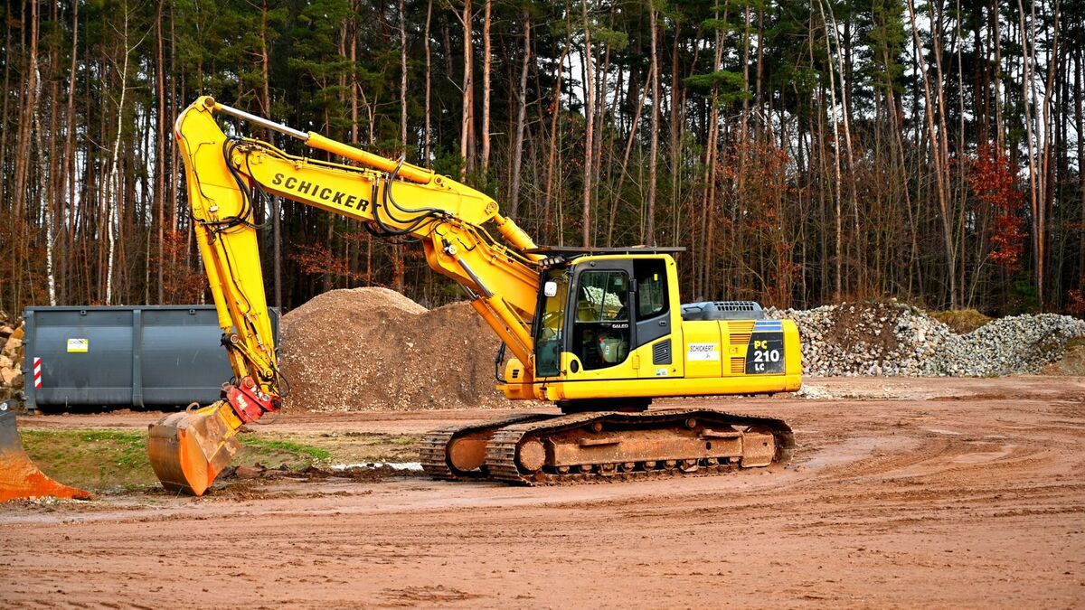 A yellow excavator working on cleared red clay ground at the edge of a stand of pine trees